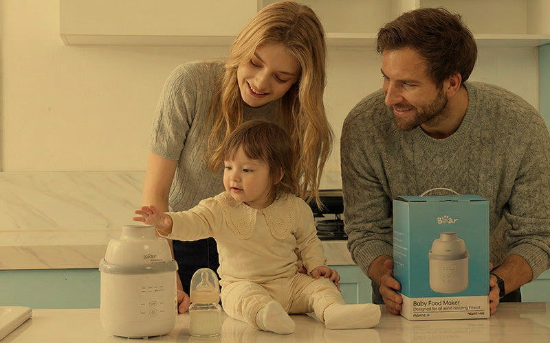 Toddler reaching for the Bear Baby Food Maker beside parents holding the box in a kitchen.