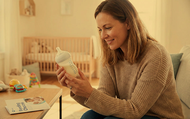 Smiling mother holding a baby milk bottle sitting on a nursery sofa near a wooden crib.