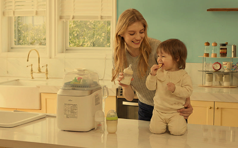 Mother holding a bottle next to her snacking toddler and a Bear Bottle Washer in a kitchen.