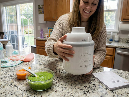 Smiling woman using the white Bear Baby Food Maker on a kitchen island next to bowls of fresh green puree.