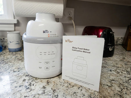White Bear Baby Food Maker sitting on a granite kitchen counter next to its printed instruction manual.