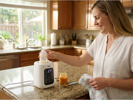 Smiling woman holding a clean glass baby bottle next to the Bear Bottle Washer Pro in a light blue kitchen.