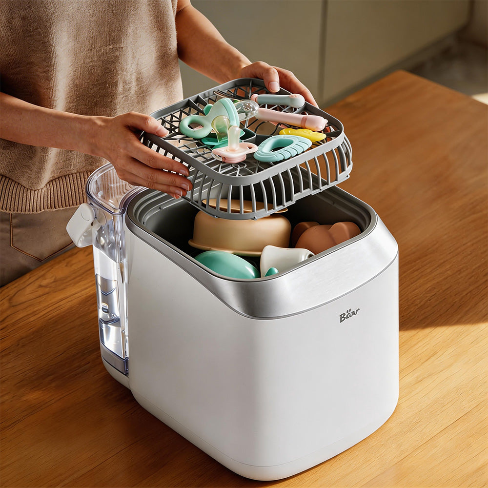 Woman loading an upper rack of pacifiers and teethers into the white Bear Bottle Washer Pro on a wooden table.